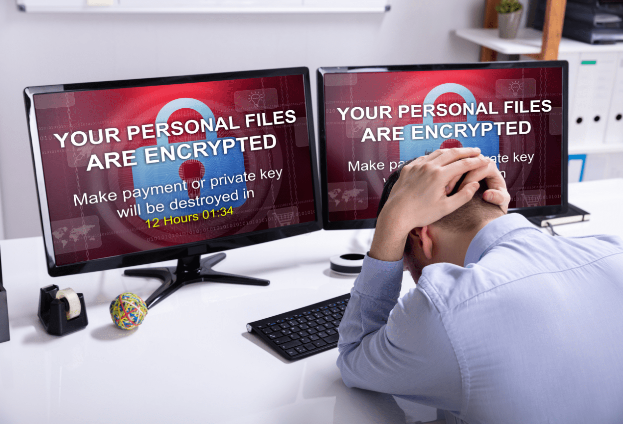 Stressed employee sitting at a desk with two monitors displaying a ransomware warning message, showing what ransomware looks like after a ransomware attack often triggered by a phishing email.